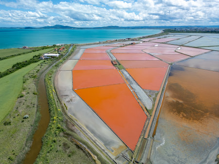 Aerial view of colorful salt pans near Burgas, Bulgaria. Vibrant red and orange evaporation ponds contrast with white salt piles and turquoise sea. Industrial and natural beauty combinedの写真素材