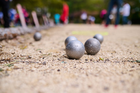Close-up of metal petanque balls on a gravel court during a game, with blurred players and colorful surroundings in the backgroundの写真素材