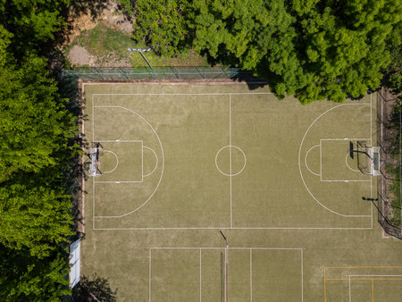 Aerial top-down view of an outdoor basketball and sports court surrounded by green trees. Empty field with visible markings and shadows on a sunny day. Recreational and athletic settingの写真素材
