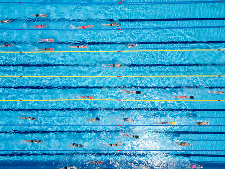 Top-down aerial view of swimmers training in an Olympic-size pool. Multiple lanes filled with people in motion. Active summer sports and fitness scene with clear blue waterの写真素材