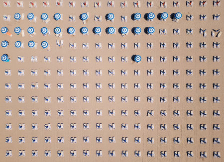 Aerial top-down view of perfectly aligned beach loungers and umbrellas on sandy ground. Repetitive geometric pattern creates a minimalist and organized summer vacation sceneの写真素材