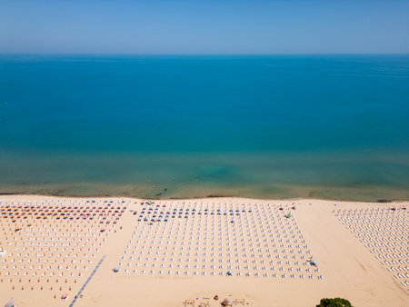 Aerial view of a wide sandy beach with rows of sunbeds and umbrellas by the calm blue sea. Symmetrical summer vacation scene with relaxing seaside atmosphere and clear skyの写真素材