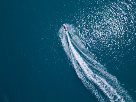 Aerial view of a jet ski speeding across dark blue water, leaving a dynamic white trail. Captured by drone from above, showcasing motion, power, and summer fun on the seaの写真素材