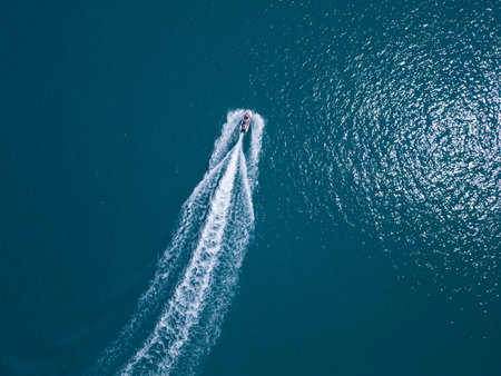 Aerial view of a jet ski speeding across dark blue water, leaving a dynamic white trail. Captured by drone from above, showcasing motion, power, and summer fun on the seaの写真素材