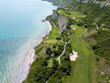 Scenic aerial view of a coastal golf course with green fairways, sand bunkers, golf carts, and players, surrounded by lush vegetation and turquoise seaの写真素材