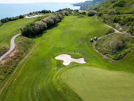 Scenic aerial view of a coastal golf course with green fairways, sand bunkers, golf carts, and players, surrounded by lush vegetation and turquoise seaの写真素材