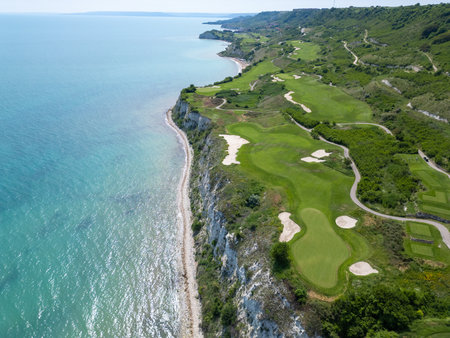 Scenic aerial view of a coastal golf course with green fairways, sand bunkers, golf carts, and players, surrounded by lush vegetation and turquoise seaの写真素材
