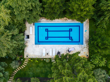 Top-down aerial view of a rectangular swimming pool surrounded by lush greenery and pine trees. Peaceful private garden retreat with sunbeds and stone pathwayの写真素材