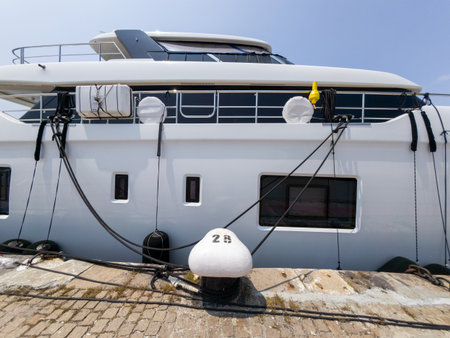Modern luxury yacht moored at a harbor dock on a sunny day. Large white vessel with black fenders and ropes, port infrastructure visible in the backgroundの写真素材