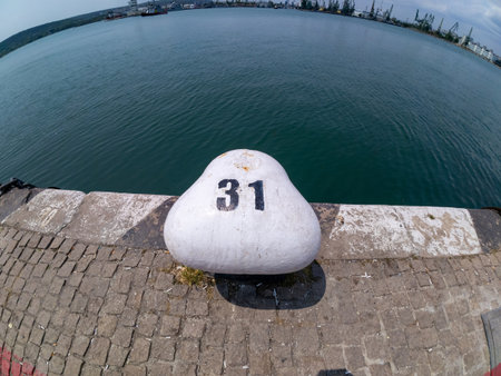 Close-up of a white mooring bollard with number 31 at a harbor dock. Calm blue sea in the background, industrial port visible across the water under a partly cloudy skyの写真素材