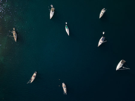 Aerial top-down view of several sailboats anchored in calm dark blue sea water with sun reflections. Peaceful summer maritime scene ideal for travel and vacation themesの写真素材