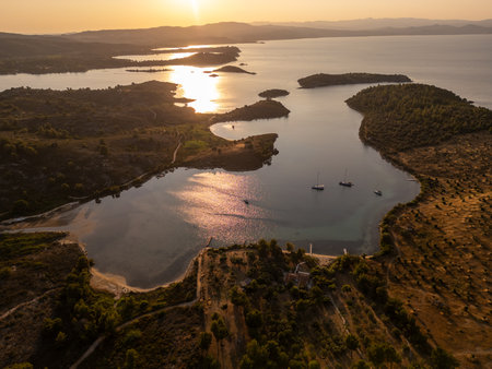 Aerial view of a serene coastal lagoon at sunset with yachts anchored near the shore, golden light reflecting on water, and hilly landscape. Peaceful Mediterranean evening sceneの写真素材