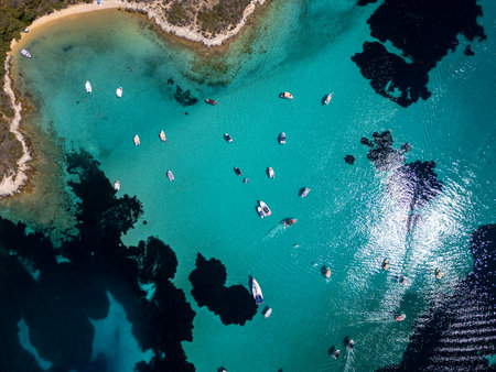 Top-down aerial view of boats floating in crystal-clear turquoise water near a sandy beach and rocky coast, with vivid sun reflections and dark underwater patchesの写真素材