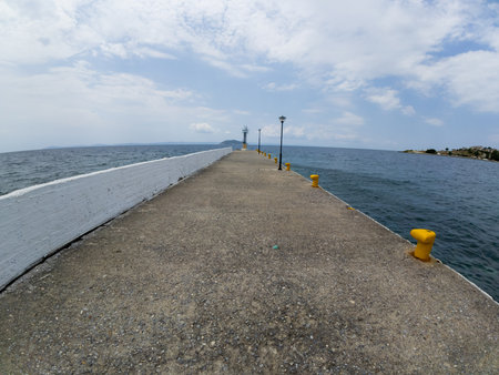 Long concrete pier with yellow mooring bollards and lampposts extending into the calm blue sea under a partly cloudy skyの写真素材