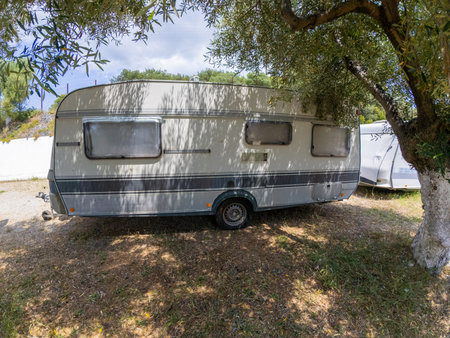 Old weathered travel trailer parked in the shade of a tree at a campsite on a sunny day, with other caravans in the backgroundの写真素材