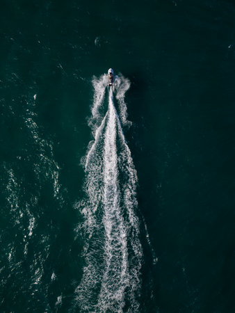 Top-down aerial view of a jet ski speeding across the deep blue sea, leaving a sharp white trail of foam. Dynamic drone footage of personal watercraft in motionの写真素材