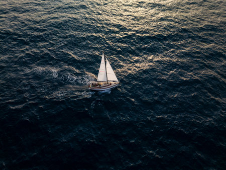 Aerial video of a white sailboat sailing alone on deep blue sea. Calm waves shimmer under sunlight. Peaceful and adventurous maritime scene perfect for travel and freedom themesの写真素材