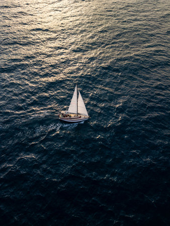 Aerial video of a white sailboat sailing alone on deep blue sea. Calm waves shimmer under sunlight. Peaceful and adventurous maritime scene perfect for travel and freedom themesの写真素材