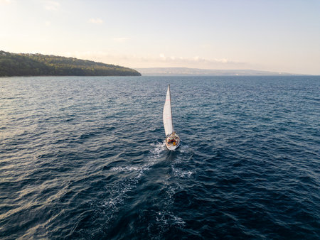 Aerial video of a sailboat cruising near a scenic coastline on a calm sea. Peaceful maritime landscape at golden hour with soft waves and gentle windの写真素材
