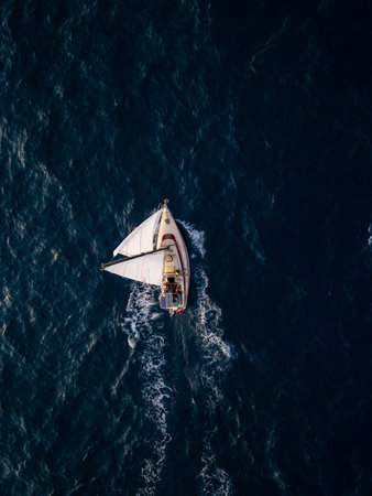 Aerial video of a white sailboat sailing alone on deep blue sea. Calm waves shimmer under sunlight. Peaceful and adventurous maritime scene perfect for travel and freedom themesの写真素材