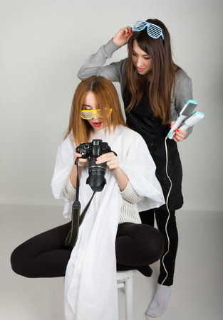 Beautiful young leggy redhaired model looks pictures on the camera, hairdresser does a hairstyle. two girls with glasses disco.の写真素材