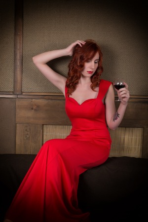 Elegant young redhead woman in a red dress, having a glass of red wine.の写真素材