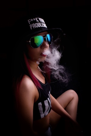 Attractive young woman in cap and sunglasses, smokes on a dark background. Be trendy, be rapper.