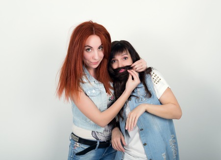 Two young girlfriends indulge and grimace, make each other a mustache out of the hair. Teenager making moustache from hair.の写真素材