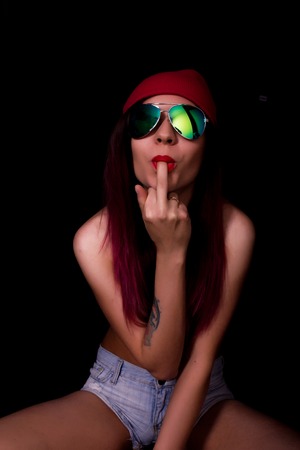 Attractive young woman in a pink knitted cap and sunglasses, sucking her middle finger on a dark background.の写真素材
