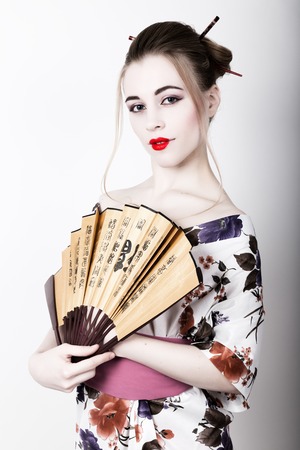 beautiful girl dressed as a geisha, she holds a chinese fan. Geisha makeup and hair dressed in a kimono. The concept of traditional Japanese values.の写真素材