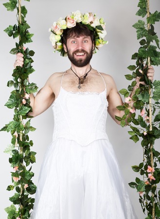 bearded man in a woman's wedding dress on her naked body, clinging to the vine. grimacing and funny. on his head a wreath of flowers. funny bearded bride.の写真素材