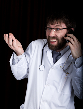 Dr. bearded manin a white medical robe, stethoscope on his neck, emotionally talking on phone.の写真素材