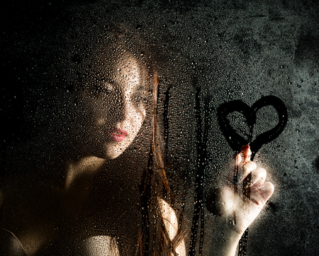 Smooth portrait of sexy model, posing behind transparent glass covered by water drops and draws a heart on the glass.の写真素材