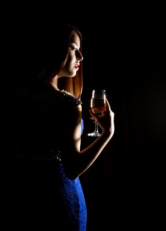 Young celebrating woman in a blue dress holding a glass of champagne on a dark background. play of light and shedow.の写真素材
