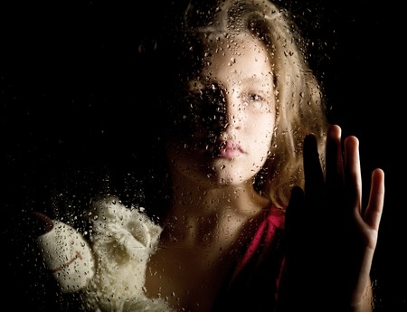 lonely young sad girl behind the window with drops, holding teddy bear and crying.の写真素材