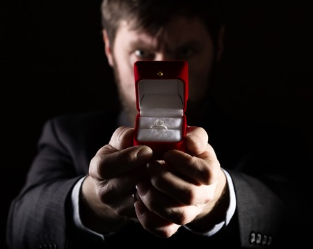 bearded man in a business suit gives a ring in the red box and expresses different emotions on a dark background.の写真素材