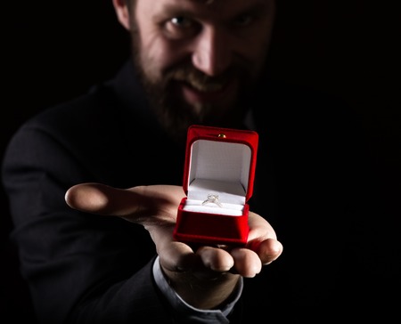 bearded man in a business suit gives a ring in the red box and expresses different emotions on a dark background.の写真素材