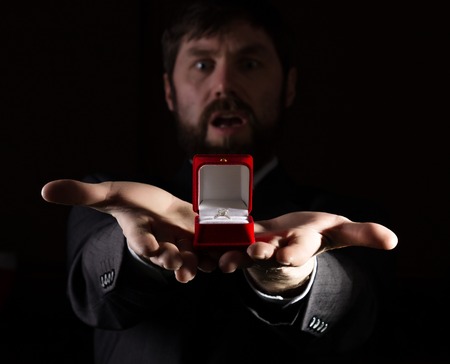 bearded man in a business suit gives a ring in the red box and expresses different emotions on a dark background.の写真素材