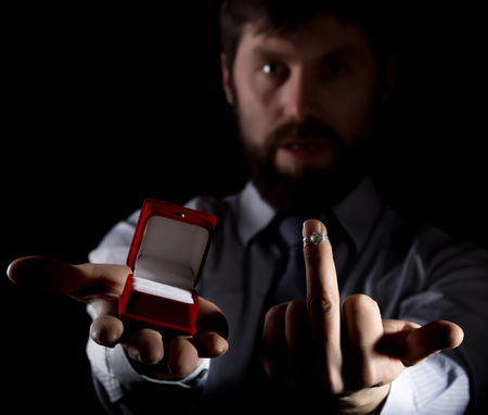 bearded man in business suit gives a ring in the red box and showing middle finger on dark...