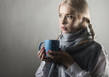 young woman dressed in sweater drinking coffee or tea, posing behind transparent glass covered by water dropsの写真素材