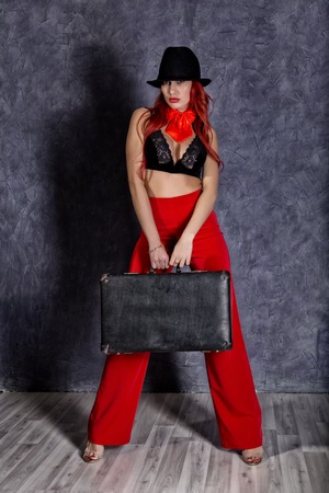 beautiful redhead woman in red pants, bra and mans hat standing and holding old suitcaseの写真素材