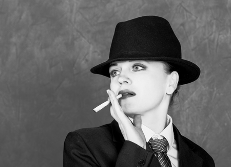Young woman in manly style with mini cigar on gray background, girl in mans suit and tie, white shirt and hatの写真素材