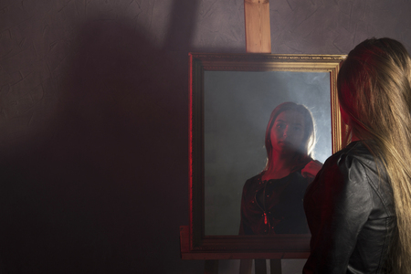 beautiful young woman in a leather jacket looks in the mirror as her portraitの写真素材