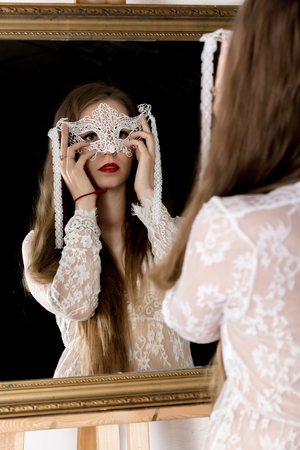 beauty young woman holding a lace mask and looks in the mirror as her portraitの写真素材