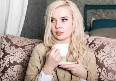Portrait of charming girl in sweater sitting on a sofa and drinking tea or coffeの写真素材