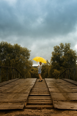 Beautiful woman hold yellow umbrella and walking on a old bridge in the countryside under rainの写真素材
