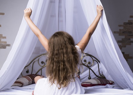 Young girl outstretching her arms sitting on the bed after good night sleep, back viewの写真素材