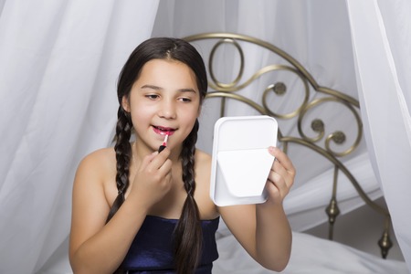 little girl sitting on a bed looks in the mirror and painting lips like her momの写真素材