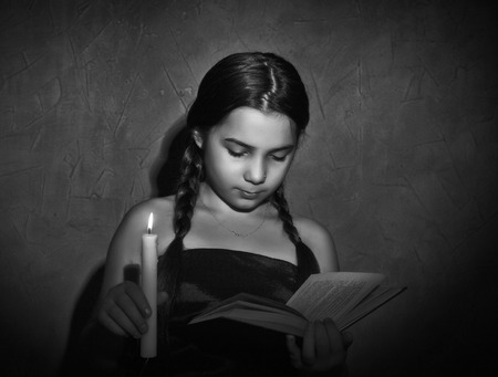 little girl reading book and studying in candle light in dark roomの写真素材