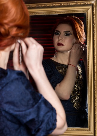 young woman puts on beautiful earrings in front of mirror. beautiful girl in vintage style.の写真素材